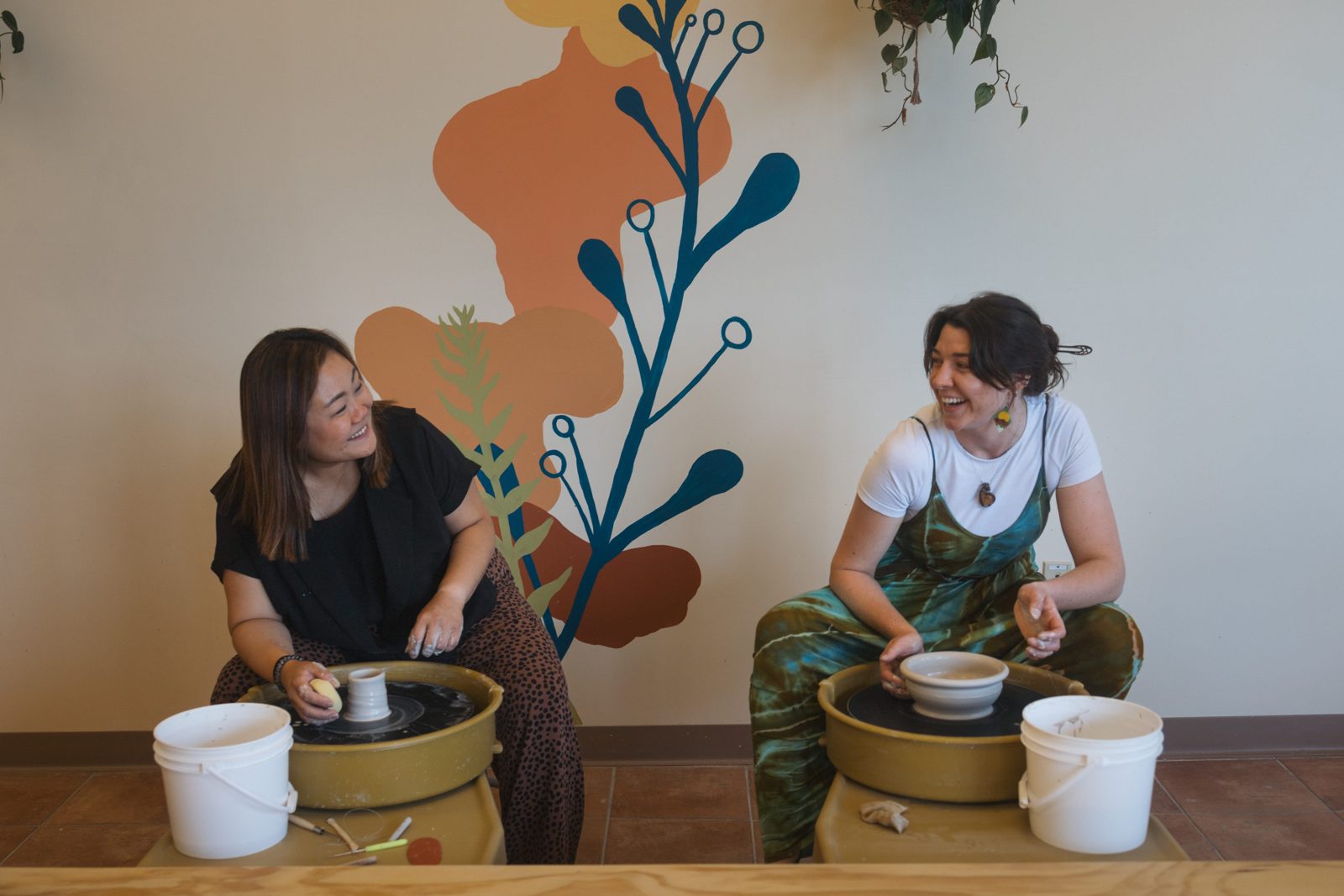 Two friends laughing together at pottery wheels in our studio, in front of the colorful Create Me Pottery mural